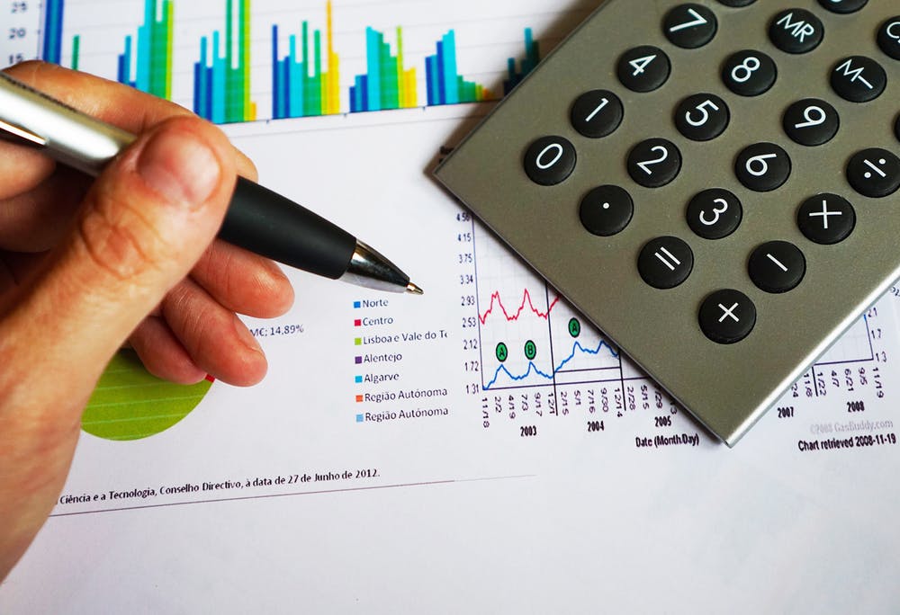 A close up of a hand holding a pen next to a calculator on top of an assortment of printed charts and graphs.